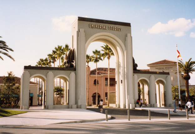 Eingang zu Universal Studios Hollywood mit einem Bogen, einer Statue, Menschen, Bäumen, Gebäuden, einem Fahnenmast und einem bewölkten Himmel.