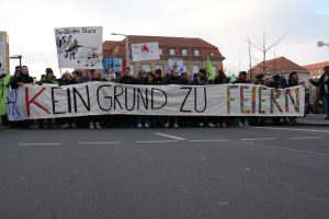 Eine Gruppe von Menschen mit einem Transparent mit der Aufschrift "Kein Grund zu Feiern" protestiert auf einer Straße mit Gebäuden, Bäumen und einem klaren Himmel im Hintergrund.