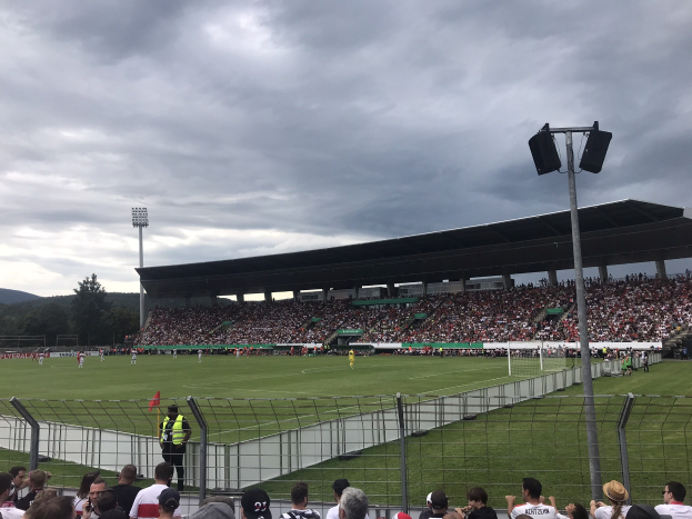 Große Zuschauermenge bei einem Fußballspiel in einem bewölkten, offenen Stadion mit beleuchtetem Feld, eingezäuntem Rand und Hügeln im Hintergrund.