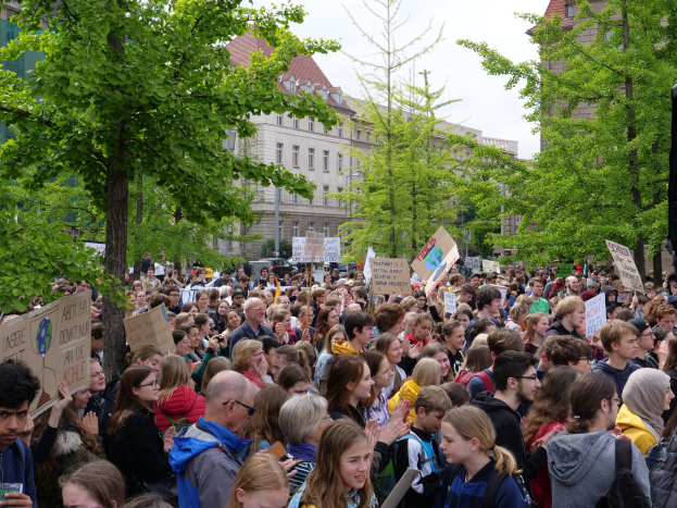 Große Menschenmenge protestiert vor einem Gebäude in Berlin, hält Schilder hoch, mit Bäumen, Fahrzeugen und einem Lautsprecher im Bild.