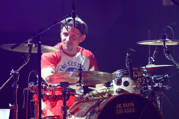 Ein Mann in einem roten und weißen T-Shirt spielt Schlagzeug auf der Bühne während eines Konzerts und lächelt, umgeben von Mikrofonen.