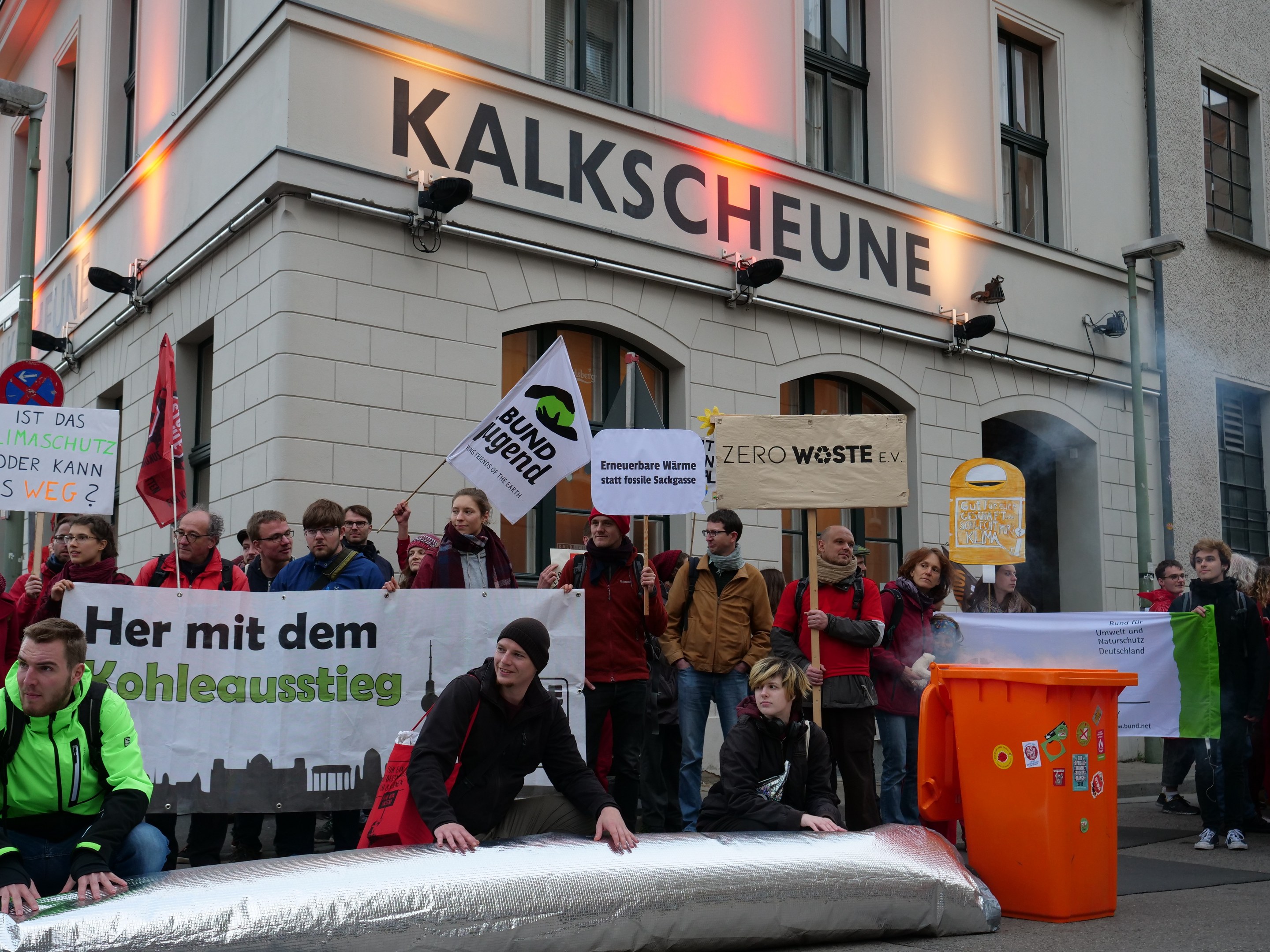 Eine Gruppe von Menschen, die vor einem Gebäude in Deutschland steht und Schilder und Plakate hält, mit zwei Personen im Vordergrund und einem Mülleimer auf der rechten Seite.