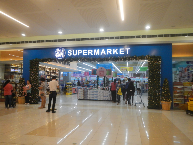 Eine Gruppe von Menschen, die vor einem Supermarkt stehen, einige halten Einkaufstaschen, mit einem Einkaufswagen, Topfpflanze, Gestellen mit Gegenständen, Schildern und einer Decke mit Lampen an der Decke.