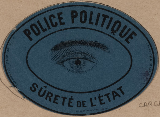 Brauner Karton mit einem blauen Aufkleber, auf dem der Text 'Polizei Politique Sûreté de l'État' und ein Auge-Symbol in der Mitte steht.