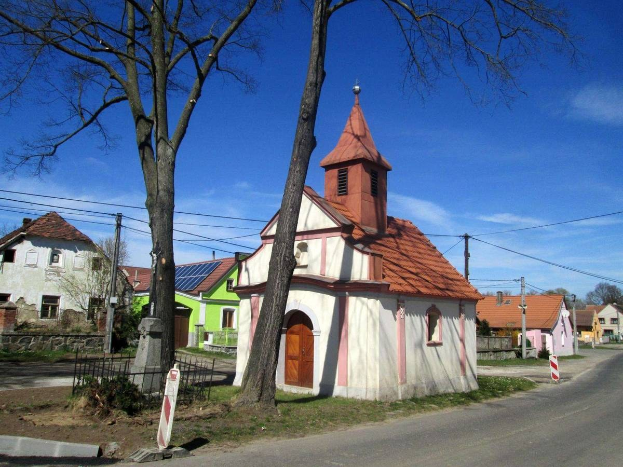 Kleine Kirche mit einem Seitenturm neben einer Straße, umgeben von Häusern, einem Metallzaun, Schildern, Gras, Pflanzen, Bäumen, Strommasten mit Drähten und einem bewölkten Himmel.