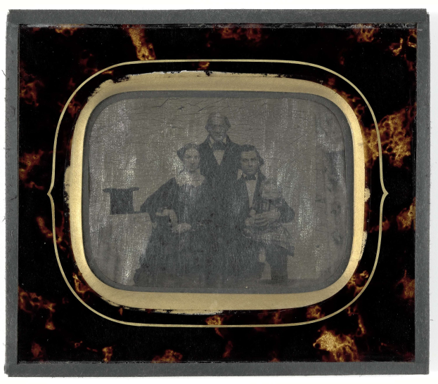 Ein Schwarz-Weiß-Foto eines Mannes in Anzug und Krawatte und einer Frau in unbekannter Kleidung, die vor einem alten Fernseher stehen und ernst schauen.