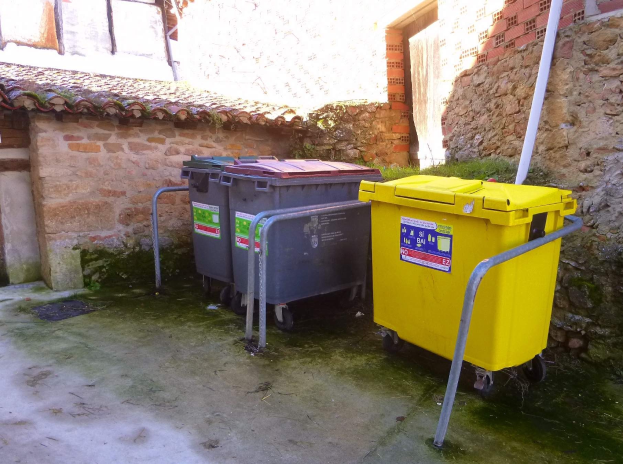 Zwei gelbe und graue Recyclingcontainer mit Rädern stehen nebeneinander vor einem Gebäude mit einer Wand und einem Fenster.