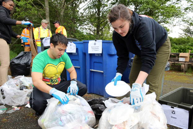 Eine Gruppe von Menschen, die Müll in einem Park sammeln, mit einem Mann und einer Frau in der Mitte, die Handschuhe tragen und Teller halten, umgeben von Plastikabdeckungen, Flaschen und anderem Müll, ein Mülleimer und eine hölzerne Bank rechts und Bäume und einen klaren blauen Himmel im Hintergrund.