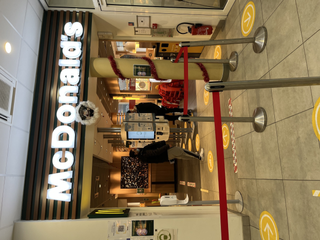 Innenansicht eines McDonald's-Restaurants mit einem sichtbaren 'McDonald's'-Schild auf der linken Seite, roten Metallstangen mit Bändern auf der rechten Seite und einigen Menschen in der Nähe des Eingangs im Hintergrund.