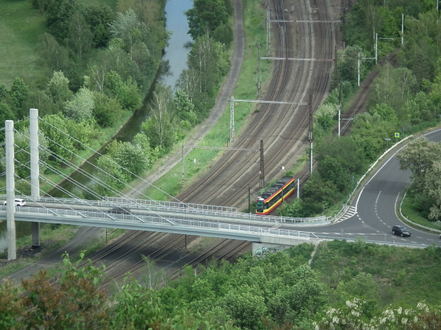 Ein Zug fährt über eine Brücke über einen Fluss, mit Fahrzeugen auf der Straße darunter und Stromleitungen entlang der Brücke, vor einem Hintergrund aus Bäumen und Gras.