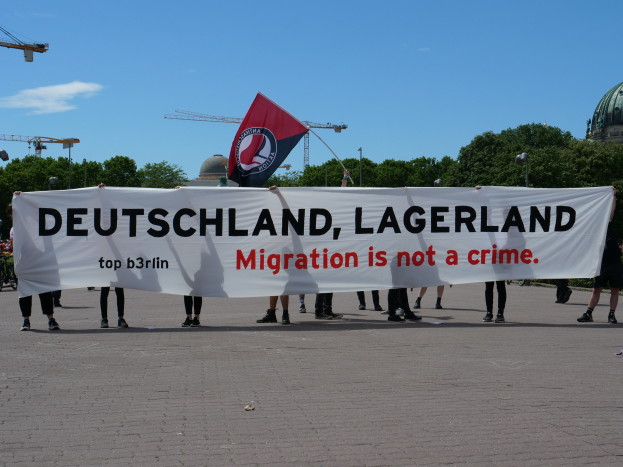 Menschen mit einem Banner, auf dem "Deutschland, Lagerland Migration ist kein Verbrechen" steht und einer Flagge, mit Straßeninfrastruktur und Gebäuden im Hintergrund.