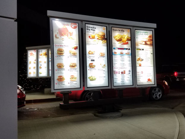 Fast Food Restaurant bei Nacht mit beleuchteter Reklame, fahrende Fahrzeuge im Hintergrund, eine Säule links und dunkler Himmel.