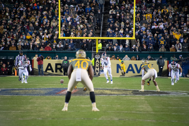 Ein Fußballspiel im Gange mit Spielern in Helmen auf dem Feld und Zuschauern in den Rängen, mit sichtbaren Pfosten und Texttafeln.