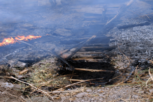 Ein Feuer lodert hell inmitten eines Schuttes, umgeben von Gras, Steinen und Holzstöcken, erhellt die Umgebung.