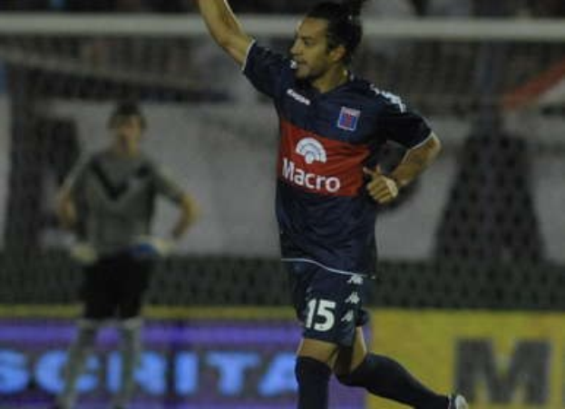 Ein Fußballer feiert auf dem Boden nach einem Tor, mit einer jubelnden Menge und einer Person im Hintergrund, alles mit einem unscharfen, energetischen Hintergrund.