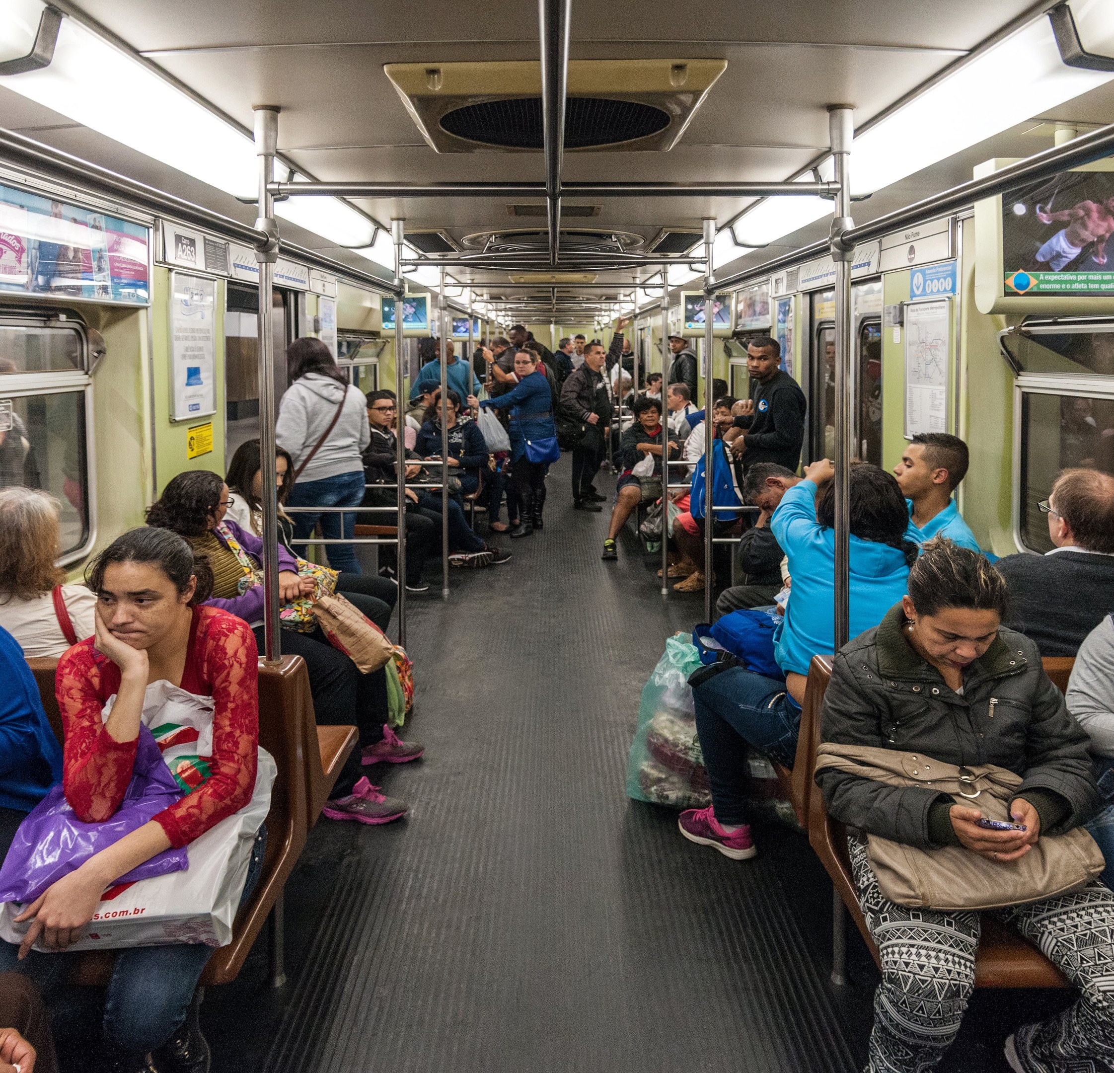 Ein überfüllter U-Bahn-Wagen mit sitzenden und stehenden Passagieren, sichtbaren Taschen, Stangen, Deckenleuchten, Wandplakaten und der Decke.