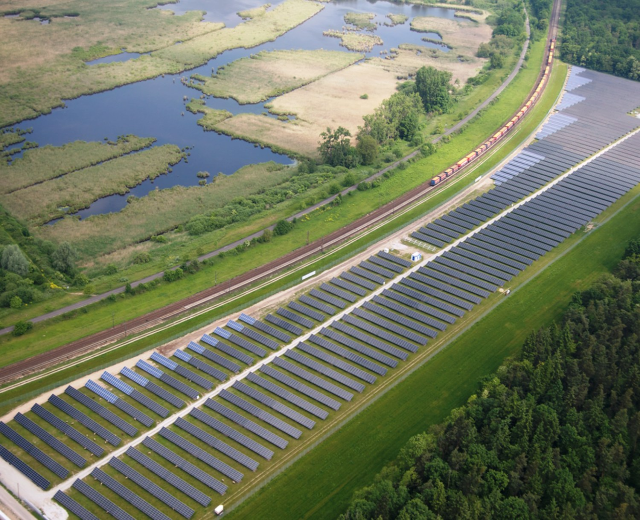 Luftaufnahme einer Solarpark mit Panelen, umgeben von Vegetation und Wasser, mit einem Zug auf einer nahen Bahnstrecke.
