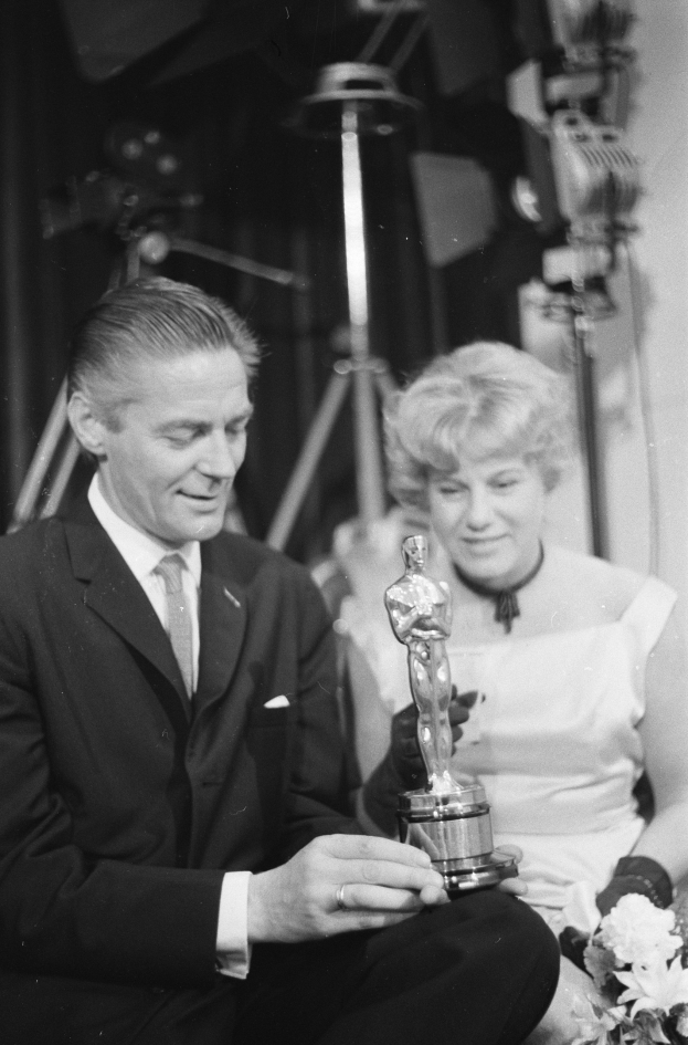 Ein Mann in einem schwarzen Blazer, weißem Hemd und Krawatte hält eine Oscar-Statue in der Hand und sitzt neben einer Frau, die einen Blumenstrauß hält, beide in eleganter Kleidung, mit Licht im Hintergrund, dargestellt in Schwarz-Weiß.