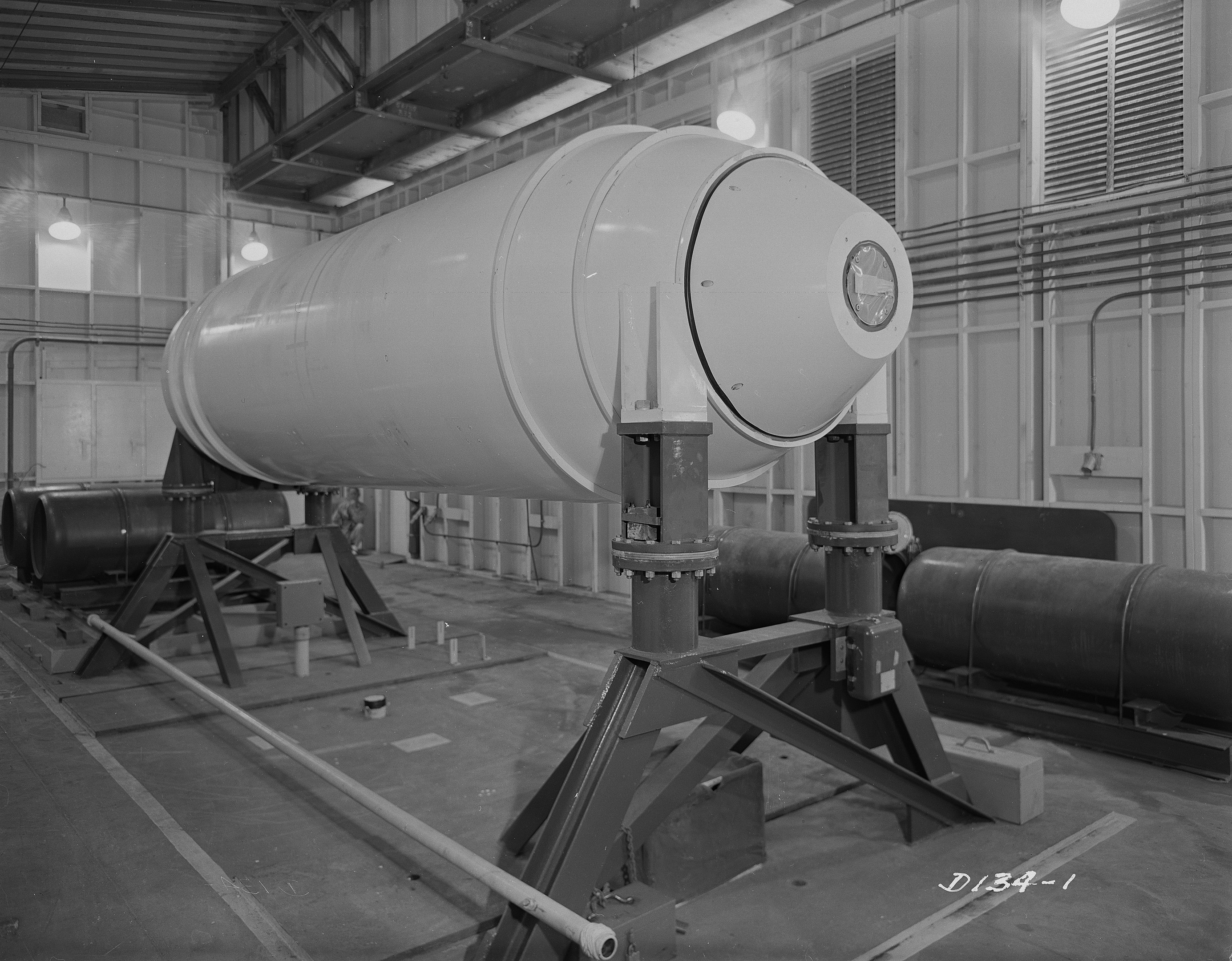 Schwarzes und weißes Foto einer großen zylindrischen Wasserstoffbombe auf einem Ständer in einer Fabrik mit Rohren, Lampen und einer Wand im Hintergrund.