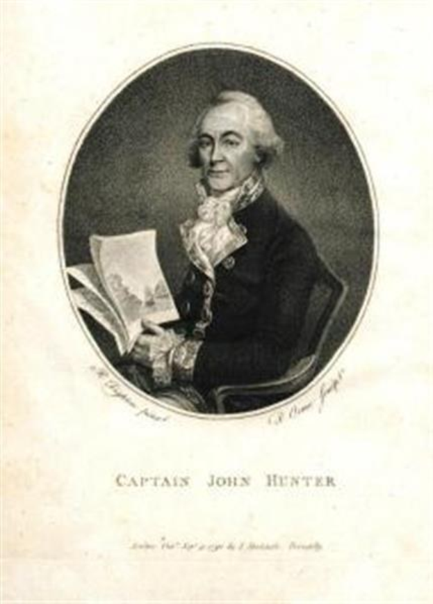 Captain John Hunter trägt einen marineblauen Anzug, ein weißes Hemd und eine rote Krawatte und sitzt in einem Stuhl, während er ein Buch liest und einen ernsten Gesichtsausdruck hat, mit dem Text "Captain John Hunter" unten.