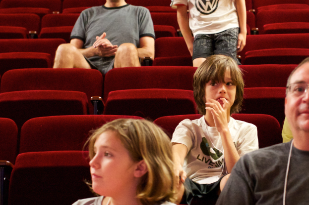Eine Gruppe von Menschen sitzt in einem Kinosaal mit roten Sesseln, wobei ein Junge in einem weißen T-Shirt und schwarzen Hosen in der Mitte steht.