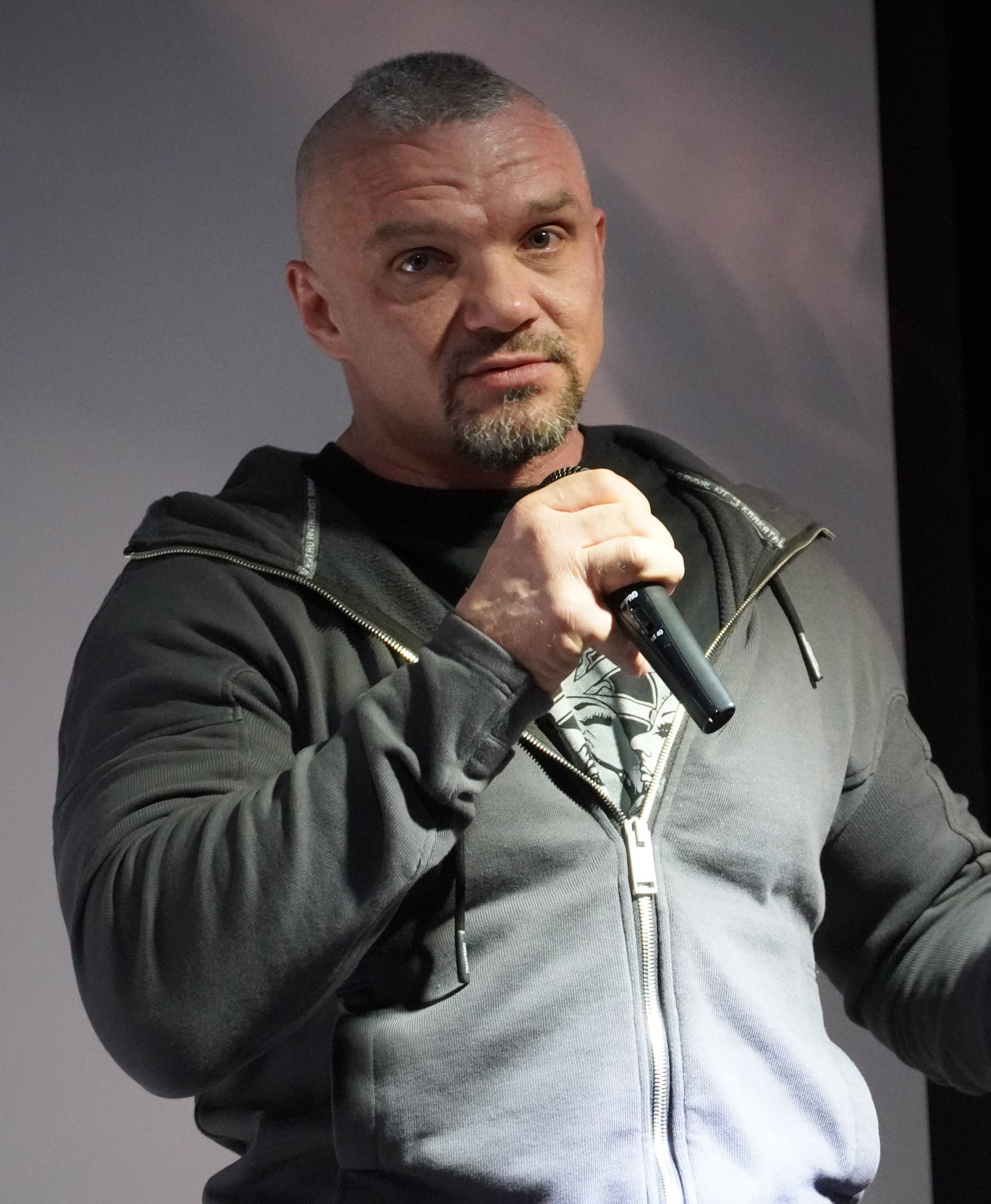 Ein Mann in einer schwarzen Kapuzenjacke und einem schwarzen Anzug mit einem wei├čen Hemd, der ein Mikrofon h├Ąlt, steht selbstbewusst vor einer Wand und scheint ein Komiker mit zurückgek├Ąmmtem Haar zu sein.