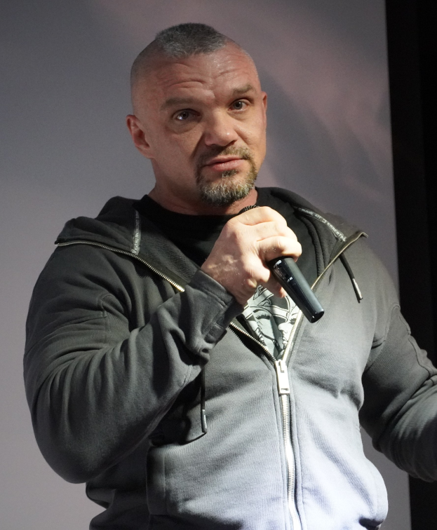 Ein Mann in einer schwarzen Kapuzenjacke und einem schwarzen Anzug mit einem wei├čen Hemd, der ein Mikrofon h├Ąlt, steht selbstbewusst vor einer Wand und scheint ein Komiker mit zurückgek├Ąmmtem Haar zu sein.