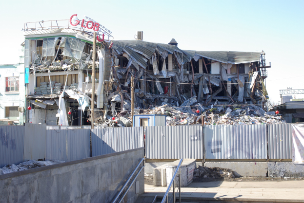 Großes Gebäude in der Mitte einer Straße abgerissen, umgeben von einem Metallzaun, mit einer Treppe, einem Schild, Pfählen, benachbarten Gebäuden, Drähten und einem bewölkten Himmel.