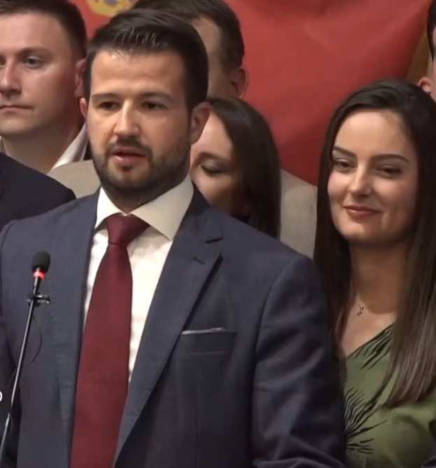 Mann in Anzug und Krawatte, der vor einer Menge an einem Rednerpult steht und eine Mikrofone hält, mit einem Banner im Hintergrund.