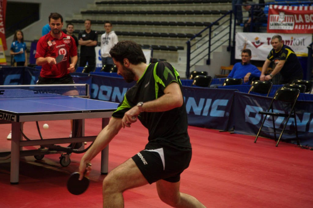 Gruppe von Menschen in verschiedenen farbigen Klamotten, die ein Spiel mit einem Ball auf einem Tisch in der Nähe von Stühlen und einer Treppe spielen.