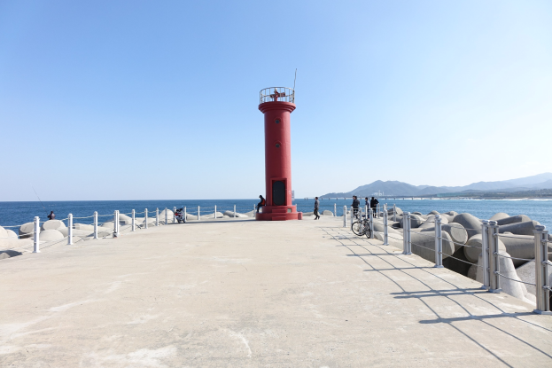 Roter Leuchtturm auf einem Pier am Meer mit Menschen, Fahrrädern, Geländern und Felsen, Hügel im Hintergrund unter einem klaren blauen Himmel.