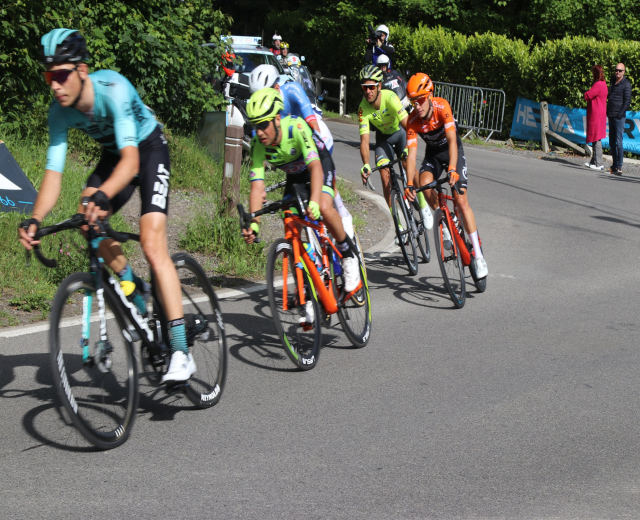 Fahrradfahrer mit Helmen und Schutzbrillen fahren auf einer Straße mit einem Auto im Hintergrund, während andere rechts stehen; es sind auch Banner, Geländer, Pflanzen und Bäume zu sehen.