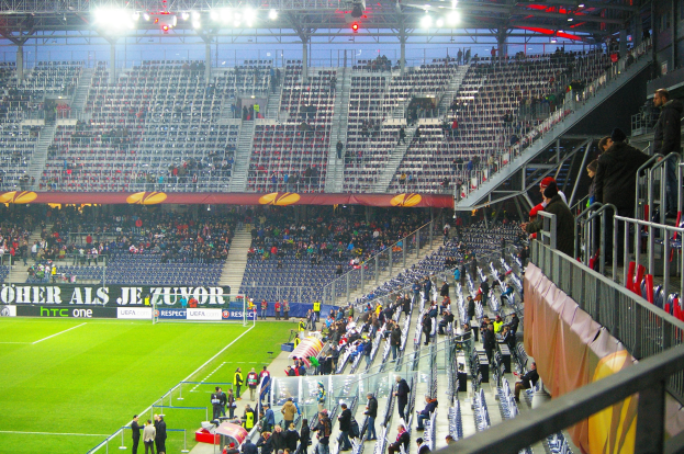 Großes Stadion mit Zuschauern bei einem Fußballspiel, einige stehen und sitzen auf Stühlen, ein Geländer und eine Fahne auf der rechten Seite und ein leerer Abschnitt mit Deckenbeleuchtung im Hintergrund.