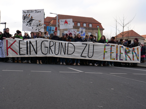 Demonstranten mit einem Banner "Kein Grund zu Feiern" gegen deutsche Sparmaßnahmen, im Hintergrund Straßeninfrastruktur und Gebäude.