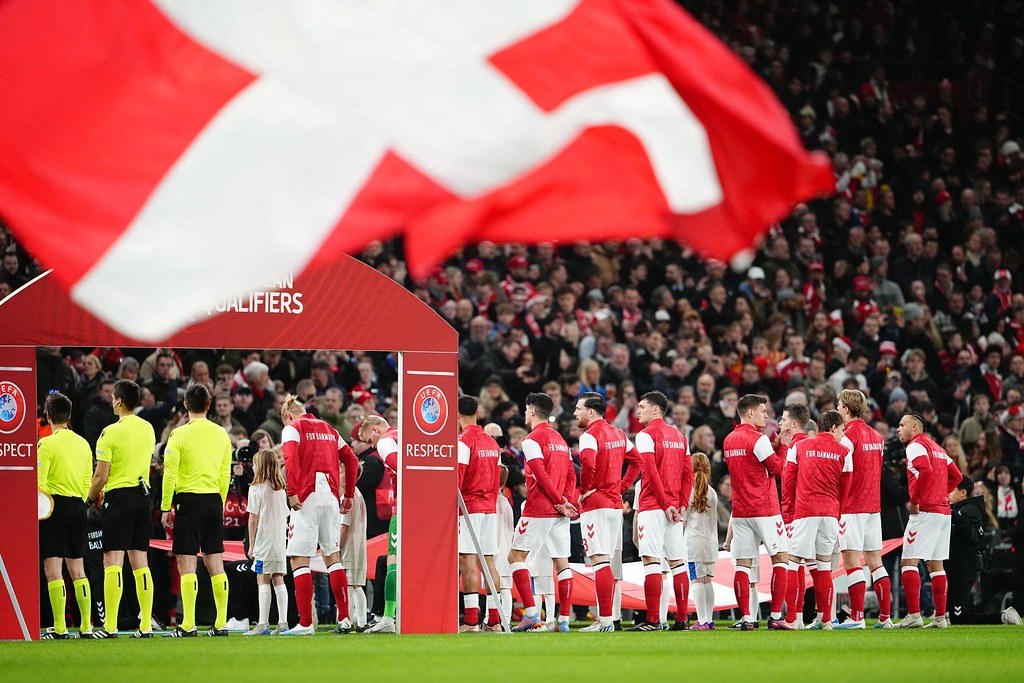 Gruppe von Menschen auf einem Fußballfeld mit einer roten und weißen Flagge im Vordergrund, einem Bogen mit der Aufschrift "Bayern München vs. Bayern München Betting Tips & Preview" im Hintergrund und einer großen Menge im Stadion.