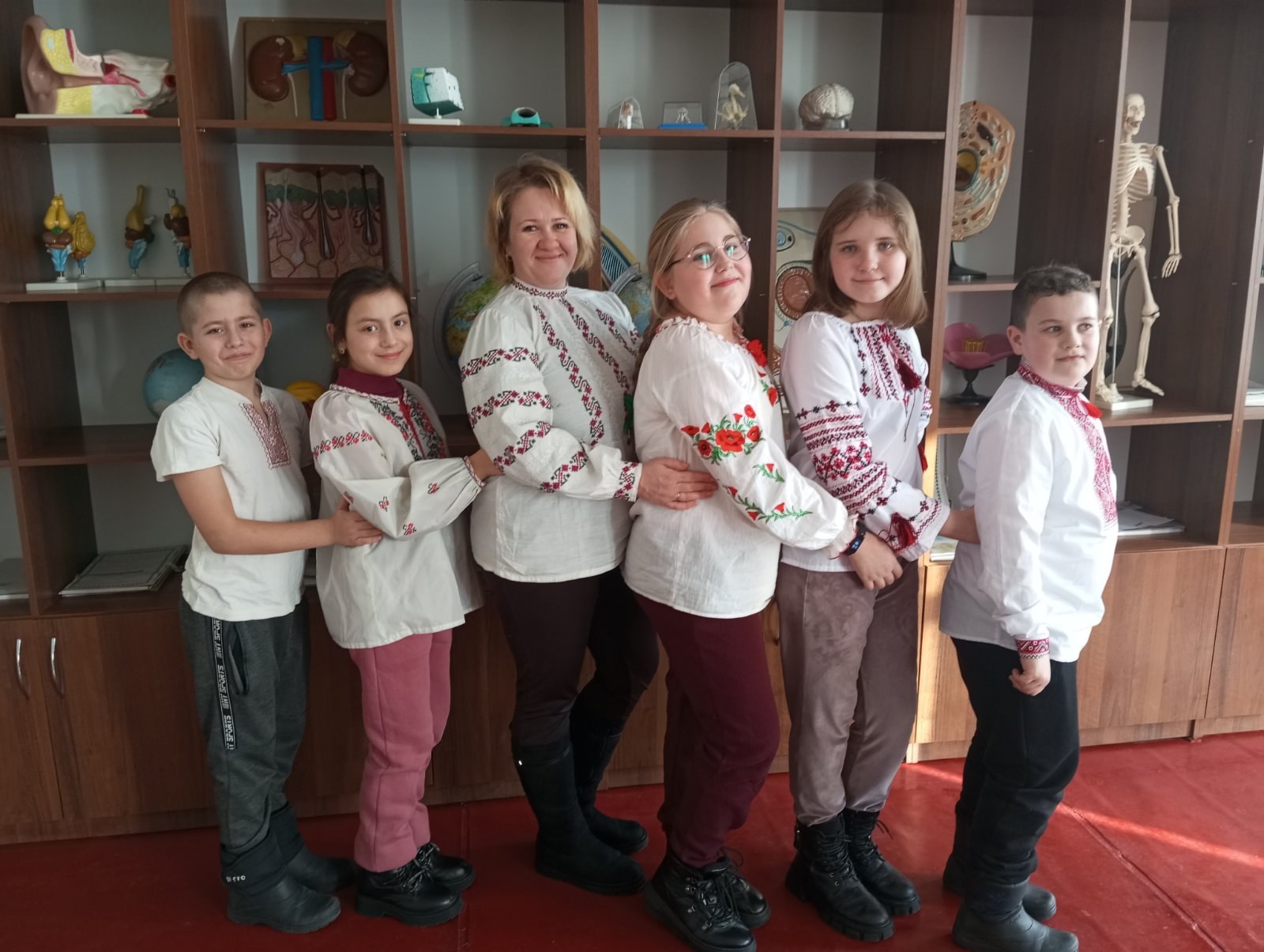 Gruppe von Kindern in traditioneller ukrainischer Kleidung vor einem Bücherregal mit Spielzeugen, einem Skelett und anderen Gegenständen.