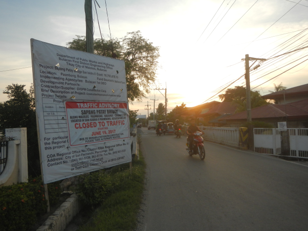 Eine Gruppe von Motorradfahrern auf einer Straße mit einem "Verkehrshinweis Geschlossen für den Verkehr"-Schild, eingerahmt von einer blühenden Wand, mit Häusern, Bäumen und einem klaren Himmel im Hintergrund.