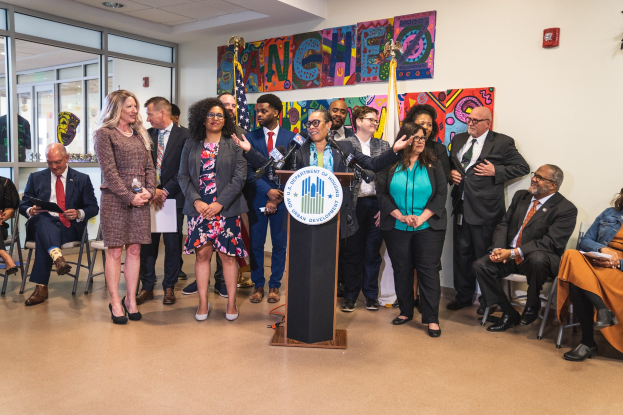 Gruppe von Menschen vor einem Podium mit einem Mikrofon und einer Tafel mit der Aufschrift "Sancho Stadtrat joins City Council", mit sitzenden und stehenden Teilnehmern, Fahnen, Feuerlöscher und Deckenleuchten im Hintergrund.
