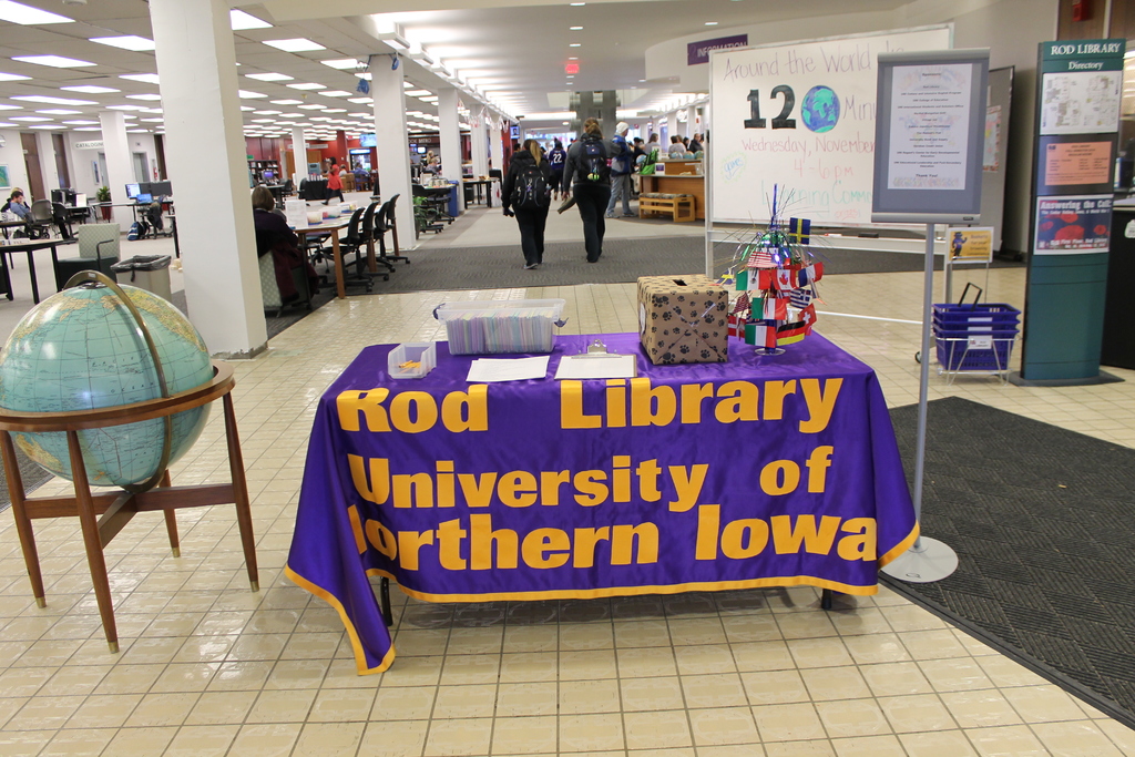 Eine Ausstellungssituation mit einem Tisch, auf dem verschiedene Landesflaggen, eine Box, Papiere und ein Banner sowie links daneben eine Weltkugel, rechts daneben ein Anschlagbrett mit einigen Menschen in der Nähe und mehrere Tische und Stühle im Hintergrund zu sehen sind.