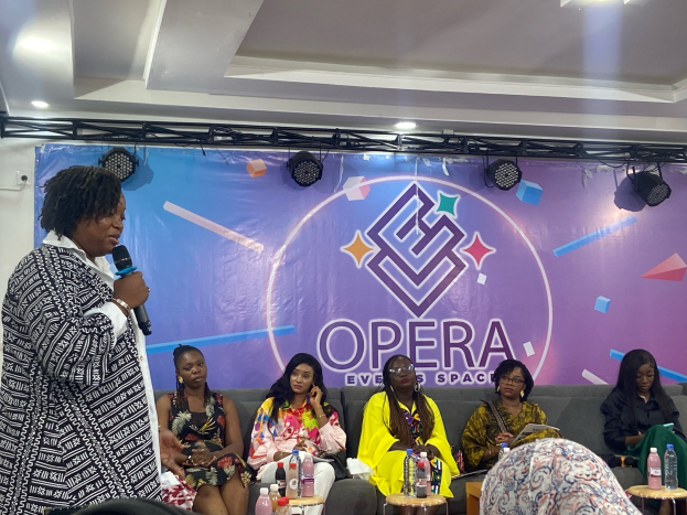 Frau mit Mikrofon steht vor sitzenden Frauen auf einer Couch bei einem Event in einem Raum mit Tischen, Flaschen, einem Banner und Deckenbeleuchtung.