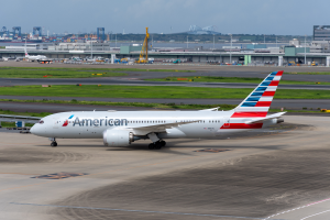 Ein Flugzeug der American Airlines steht auf der Rollbahn eines Flughafens, umgeben von Gras, Fahrzeugen, Gebäuden, Masten, Kränen und anderen Gegenständen. Im Hintergrund gibt es Bäume und einen klaren blauen Himmel.