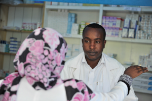 Eine Apothekerin in einem weißen Kittel spricht mit einer sitzenden Frau in einer Apotheke, mit Regalen voller Schachteln und anderen Gegenständen im Hintergrund.