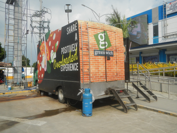 Ein Lastwagen vor einem Gebäude mit einer Tafel, auf der "Teilen Sie eine positiv überladene Erfahrung" steht, mit Stufen, einem Zylinder auf dem Boden, Geländern, Strommasten mit Drähten, Laternen, Bäumen und einem bewölkten Himmel im Hintergrund.