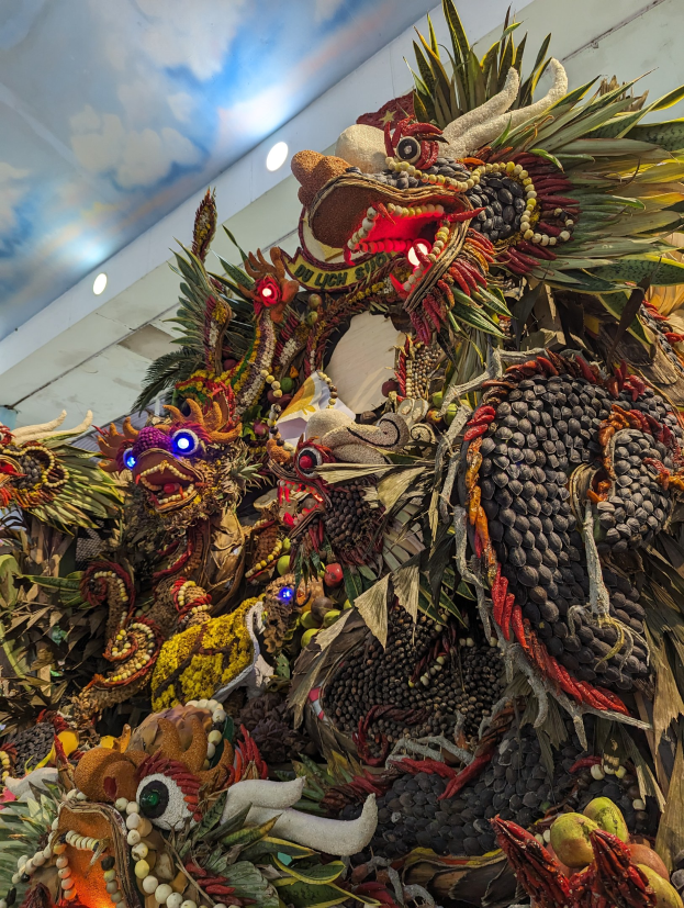 Ein großer Haufen Obst und Gemüse in einem Raum mit einer beleuchteten Decke und einer Wand im Hintergrund, mit dem Text 'Papua New Guinea Carnival' unten.