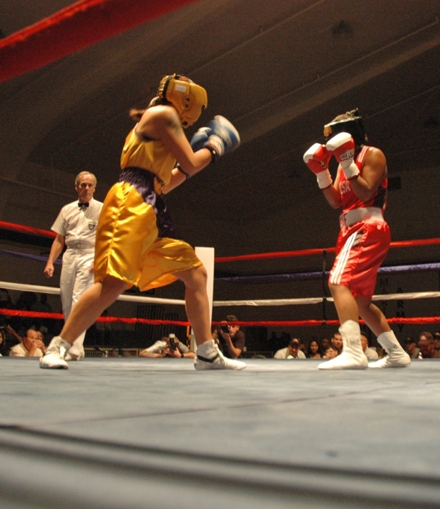 Zwei Boxer im Ring mit einem Schiedsrichter, umgeben von Fotografen und Zuschauern unter einer sichtbaren Decke.