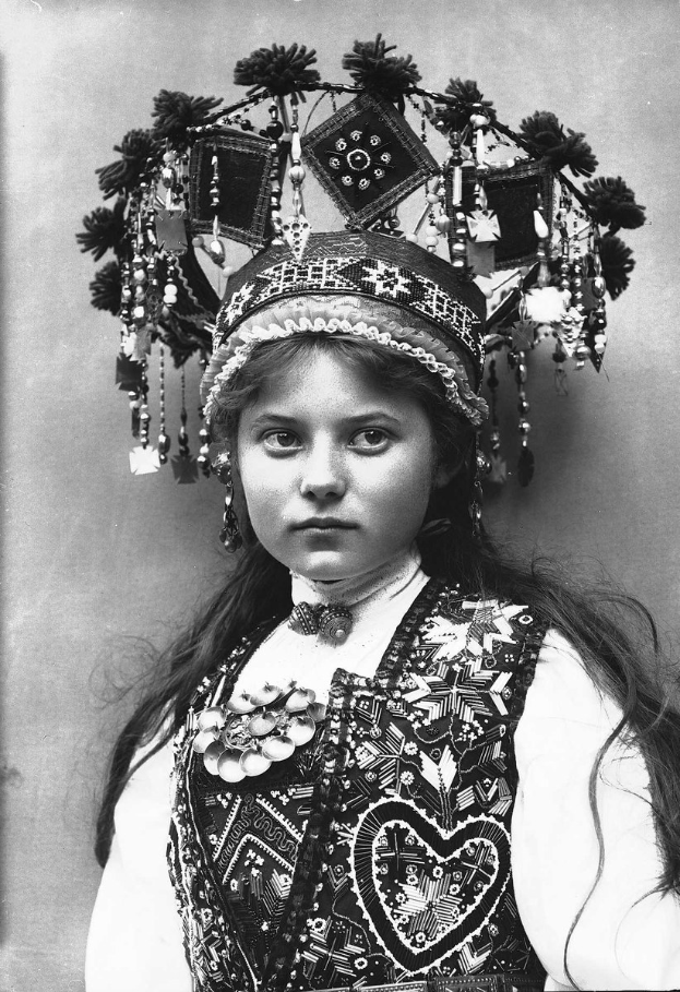 Ein Schwarz-Weiß-Foto eines jungen Mädchens in traditioneller russischer Kleidung mit besticktem Kleid, Blumenkopfschmuck und zurückgebundenem Haar.