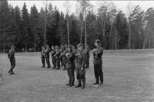 Schwarzes Bild einer Gruppe von Männern in Militäruniformen mit Mützen und Gewehren, die in einem Feld mit Bäumen und einem klaren Himmel im Hintergrund stehen, wahrscheinlich während eines Trainingsübungs für deutsche Truppen.