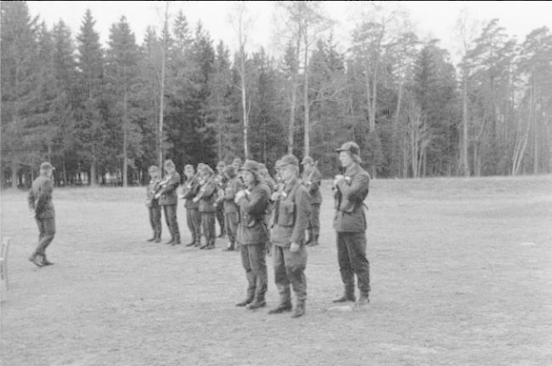 Schwarzes Bild einer Gruppe von Männern in Militäruniformen mit Mützen und Gewehren, die in einem Feld mit Bäumen und einem klaren Himmel im Hintergrund stehen, wahrscheinlich während eines Trainingsübungs für deutsche Truppen.