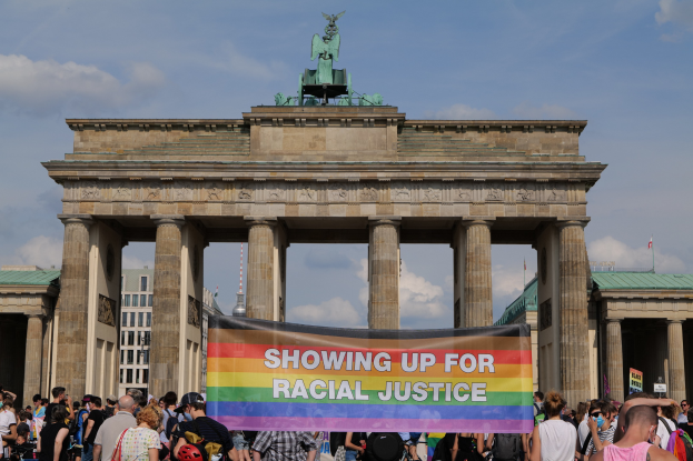 Eine Gruppe von Menschen mit einer 'Rassengerechtigkeit'-Schleife vor dem Reichstagsgebäude in Berlin, Deutschland, mit Gebäuden und einem bewölkten Himmel im Hintergrund.