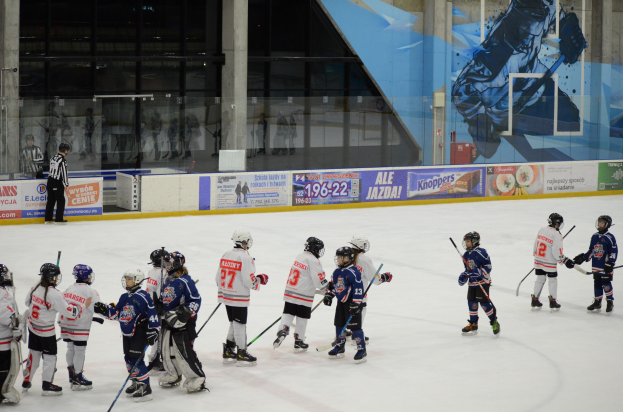 Eine Gruppe junger Menschen in Helmen und Hockey-Ausrüstung, die auf einer Indoor-Eisbahn Eishockey spielen, mit Bannern, Säulen und Glaswänden im Hintergrund.